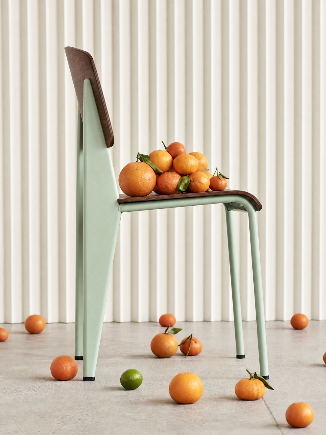 A mint-green steel and walnut chair styled with a pile of oranges against fluted cream curtains